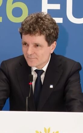Man in a black suit and tie giving a speech at a podium, with a blue backdrop that reads 'Cyprus Presidency of the Council'.