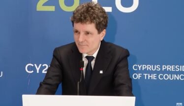 Man in a black suit and tie giving a speech at a podium, with a blue backdrop that reads 'Cyprus Presidency of the Council'.