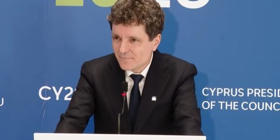 Man in a black suit and tie giving a speech at a podium, with a blue backdrop that reads 'Cyprus Presidency of the Council'.