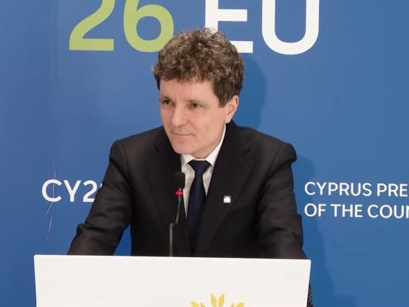 Man in a black suit and tie giving a speech at a podium, with a blue backdrop that reads 'Cyprus Presidency of the Council'.