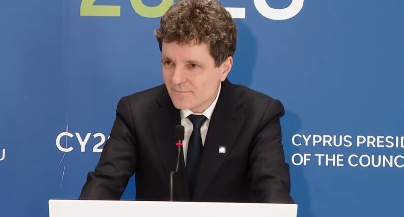 Man in a black suit and tie giving a speech at a podium, with a blue backdrop that reads 'Cyprus Presidency of the Council'.