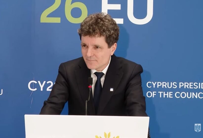 Man in a black suit and tie giving a speech at a podium, with a blue backdrop that reads 'Cyprus Presidency of the Council'.