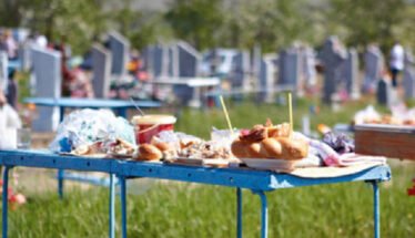 Blue folding table piled with pastries, cups, and snacks in a cemetery yard with headstones in the background.