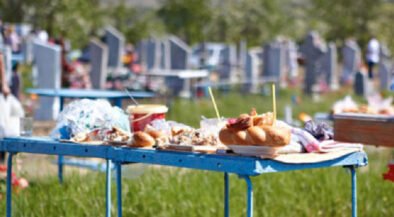 Blue folding table piled with pastries, cups, and snacks in a cemetery yard with headstones in the background.