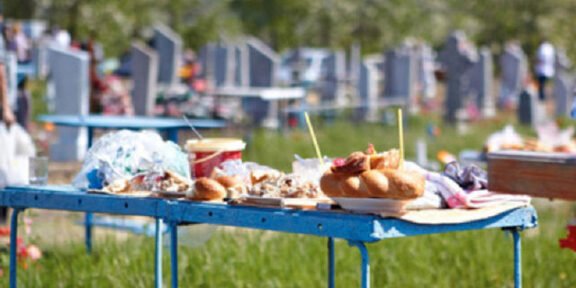 Blue folding table piled with pastries, cups, and snacks in a cemetery yard with headstones in the background.