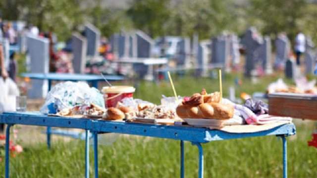 Blue folding table piled with pastries, cups, and snacks in a cemetery yard with headstones in the background.
