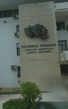 Front view of a Romanian government building with a stone pillar and emblem, the sign reading 'Guvernul României' and 'Constanța'