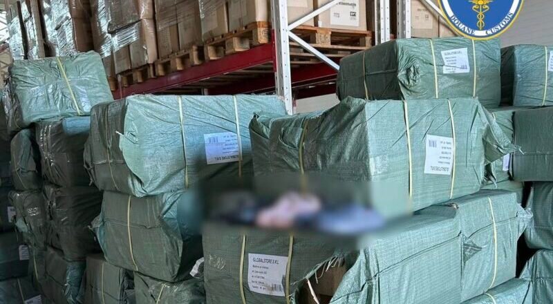 Stacks of green-wrapped bundles on wooden pallets in a warehouse, with a Romanian customs logo visible in the top right corner and labels on the bundles.
