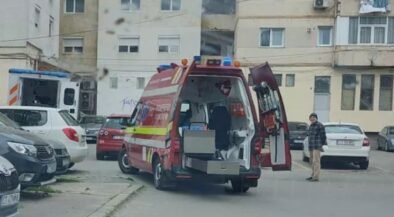 Red emergency ambulance with rear doors open, parked in a residential courtyard while a man stands nearby and a building blocks the background.