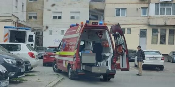 Red emergency ambulance with rear doors open, parked in a residential courtyard while a man stands nearby and a building blocks the background.