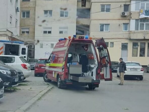 Red emergency ambulance with rear doors open, parked in a residential courtyard while a man stands nearby and a building blocks the background.
