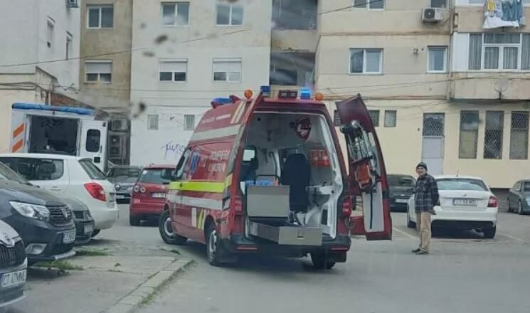 Red emergency ambulance with rear doors open, parked in a residential courtyard while a man stands nearby and a building blocks the background.