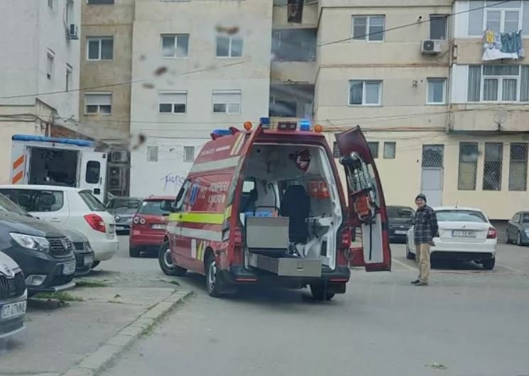 Red emergency ambulance with rear doors open, parked in a residential courtyard while a man stands nearby and a building blocks the background.
