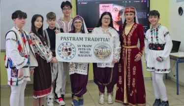 Group of students in colorful traditional outfits posing indoors, holding a banner that reads 'Pastram Traditia!' (We live in a better environment).
