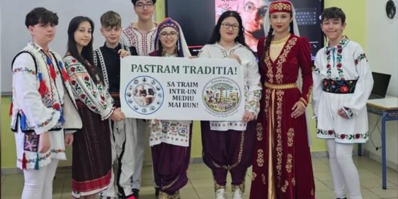 Group of students in colorful traditional outfits posing indoors, holding a banner that reads 'Pastram Traditia!' (We live in a better environment).