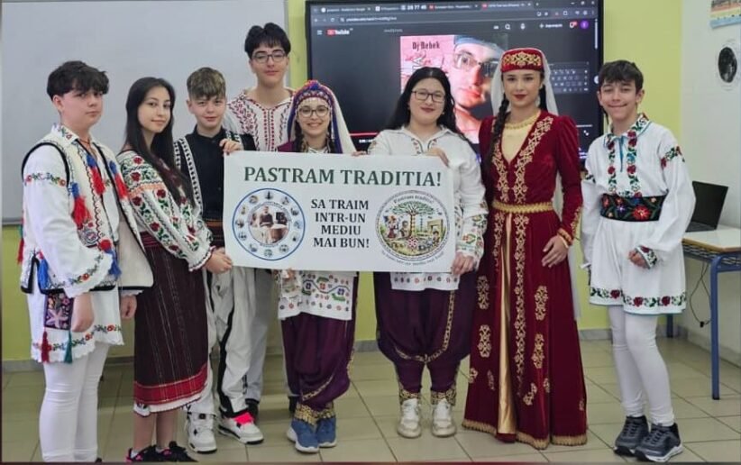 Group of students in colorful traditional outfits posing indoors, holding a banner that reads 'Pastram Traditia!' (We live in a better environment).