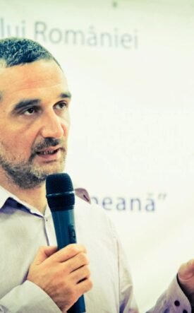 Man with short hair and beard speaks into a handheld microphone while gesturing with his right hand, standing in front of a Romanian event banner.