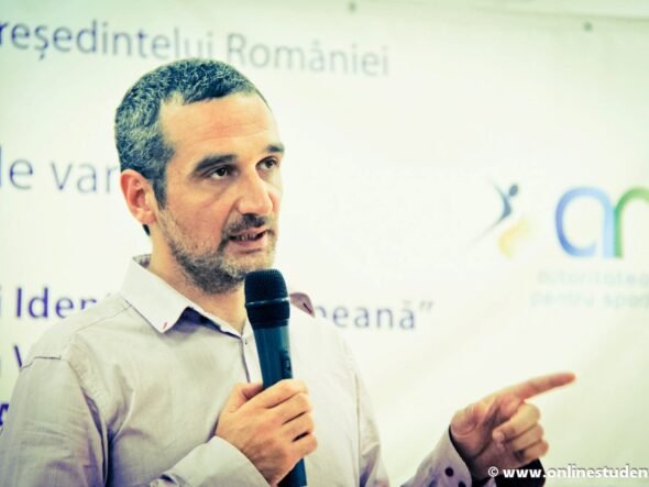 Man with short hair and beard speaks into a handheld microphone while gesturing with his right hand, standing in front of a Romanian event banner.