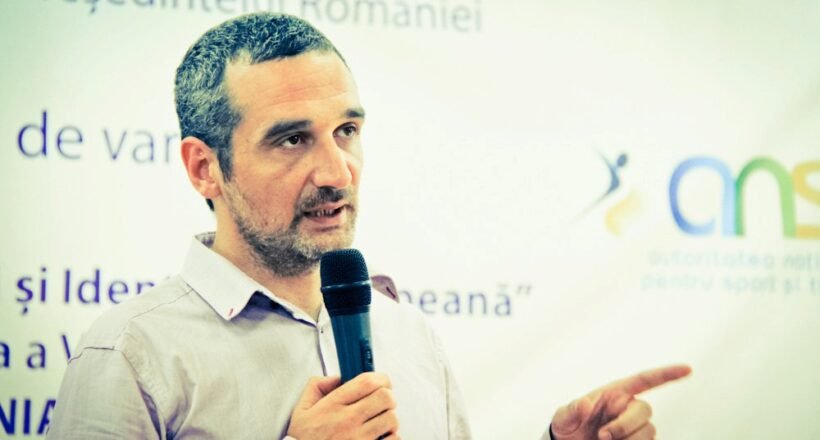 Man with short hair and beard speaks into a handheld microphone while gesturing with his right hand, standing in front of a Romanian event banner.