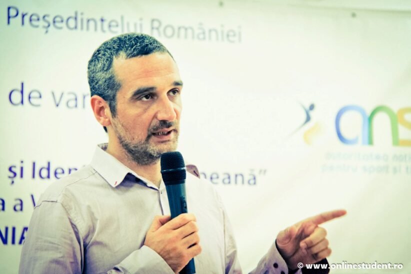 Man with short hair and beard speaks into a handheld microphone while gesturing with his right hand, standing in front of a Romanian event banner.