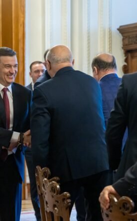Businessman in a dark suit and red tie shakes hands with another man in a formal hallway setting.