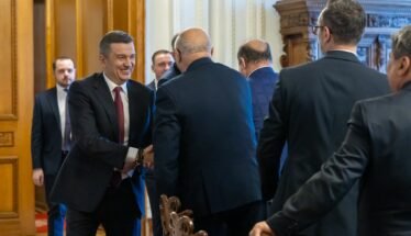 Businessman in a dark suit and red tie shakes hands with another man in a formal hallway setting.