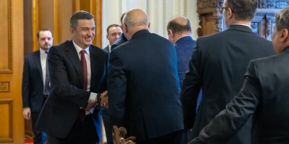 Businessman in a dark suit and red tie shakes hands with another man in a formal hallway setting.