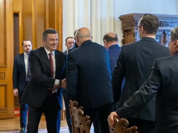 Businessman in a dark suit and red tie shakes hands with another man in a formal hallway setting.