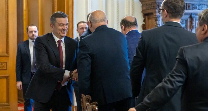 Businessman in a dark suit and red tie shakes hands with another man in a formal hallway setting.