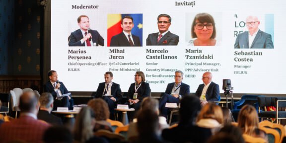 Panel discussion on PPP financing is taking place on a stage with a moderator and five panelists seated in front of a large screen showing their photos and names; audience watches from the foreground.