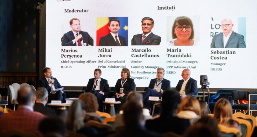 Panel discussion on PPP financing is taking place on a stage with a moderator and five panelists seated in front of a large screen showing their photos and names; audience watches from the foreground.