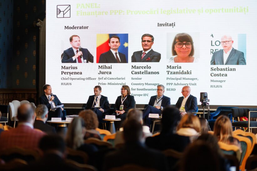Panel discussion on PPP financing is taking place on a stage with a moderator and five panelists seated in front of a large screen showing their photos and names; audience watches from the foreground.