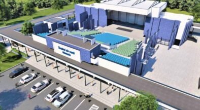 A rendered aerial view of a modern sports arena with blue and green seating, a large entrance, and a parking lot nearby.