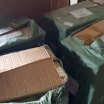 Stack of cardboard boxes wrapped in green plastic tarps, some taped and torn, stored in a corner.