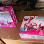 Two pink children's toy sets in colorful cartoon packaging resting on brown cardboard boxes in what looks like a storage area.