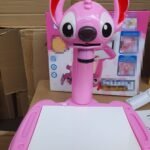 Pink kids' drawing tablet with a wide-eyed cartoon dog head on top and a white drawing surface behind it, set on a cardboard backdrop.