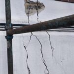 Exterior wall with large, jagged cracks and exposed sections behind rusted scaffolding pipes.