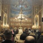 Crowded Orthodox church interior with an ornate iconostasis, frescoes, and a large chandelier overhead.