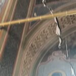 Part of an ornate religious ceiling mural showing a saint with a blue robe and halo, damaged by cracking and hanging wires.