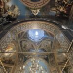 Ornate church ceiling with frescoes and arches, a blue circular window at the center, and a gold chandelier below; cracks run along the walls.