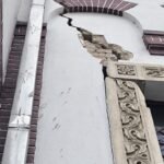 White building facade with a long vertical crack in the wall, exposed rubble, decorative carved stonework on the right, and a green pipe overhead.