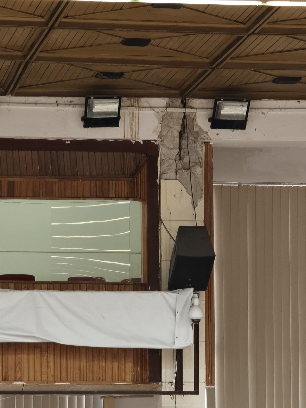 Indoor room corner showing cracked plaster on the wall beside a mounted black speaker and a wooden-framed window.