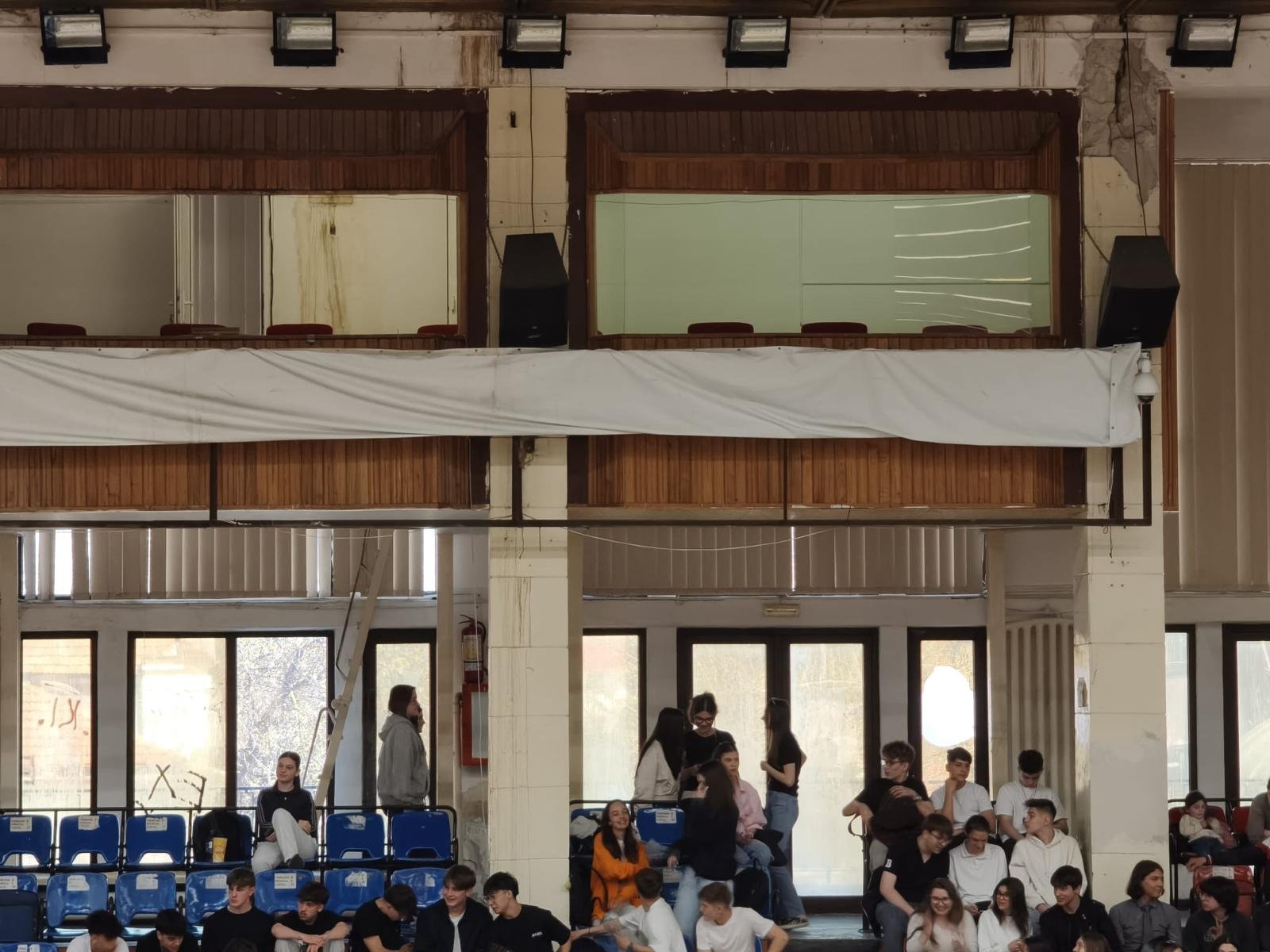 Group of students seated on blue bleachers inside a spacious hall, with a white banner across the upper level and people standing near columns.