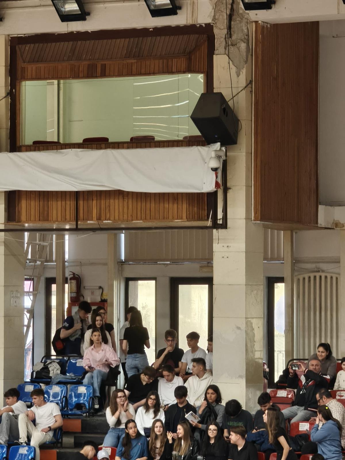 Group of students gathered in a school auditorium, some seated in blue chairs and others standing near concrete pillars and a wooden balcony above.