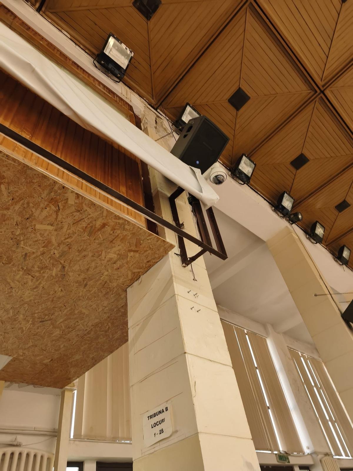 Interior of a conference hall with a wood-paneled ceiling, wall speakers and stage lights, a white column, and a sign reading 'Tribuna B Locuri'