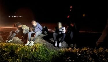 Five people in casual clothes sit on a rocky curb by a dark road at night, faces blurred, with backpacks nearby.