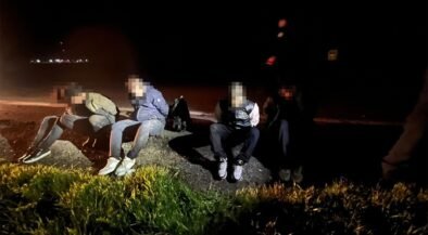 Five people in casual clothes sit on a rocky curb by a dark road at night, faces blurred, with backpacks nearby.