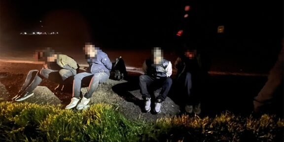 Five people in casual clothes sit on a rocky curb by a dark road at night, faces blurred, with backpacks nearby.