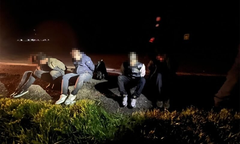 Five people in casual clothes sit on a rocky curb by a dark road at night, faces blurred, with backpacks nearby.