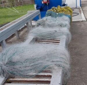 Tangled fishing nets and ropes spilled over a bench and drain near a blue industrial tank outdoors.